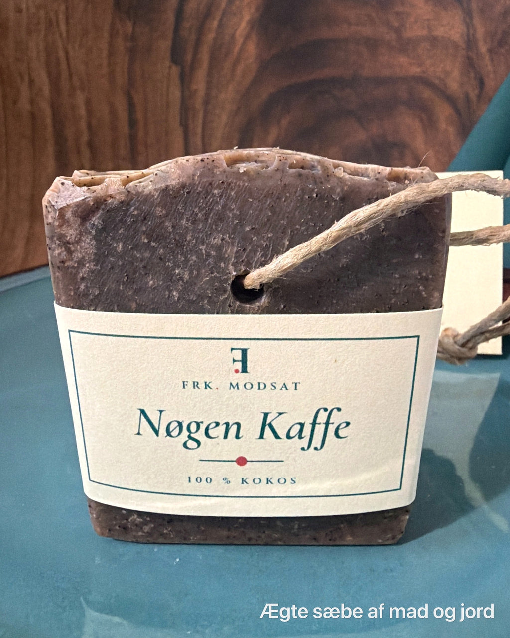 Nøgen Kaffe, mørkebrun kokossæbe med synlig kaffe, hampesnor og kartonbånd, set forfra på en teal tallerken