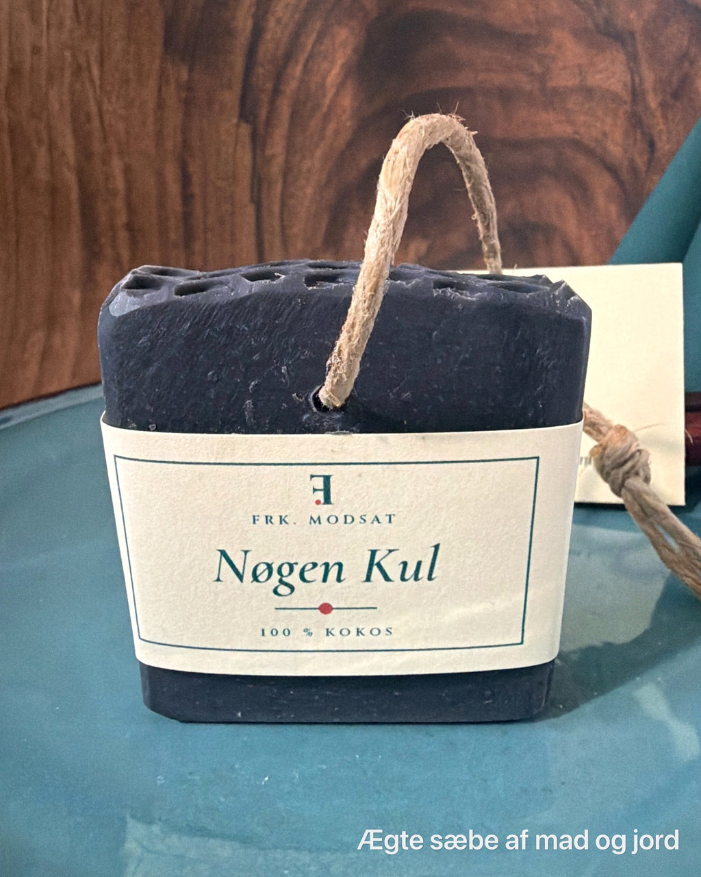 Nøgen Kul, sort kokossæbe med hampesnor og kartonbånd, set forfra på en teal tallerken